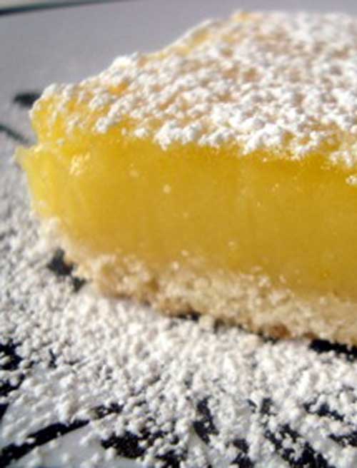 A square lemon bar on a white plate. The top of the bar and the plate have been sprinkled with powdered sugar.