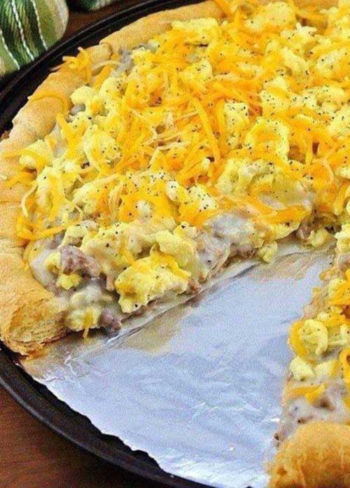 A finished Sausage Gravy Breakfast Pizza rests on a round, foil lined pan; showing of the sausage, gravy, eggs, and cheese. One piece is missing.