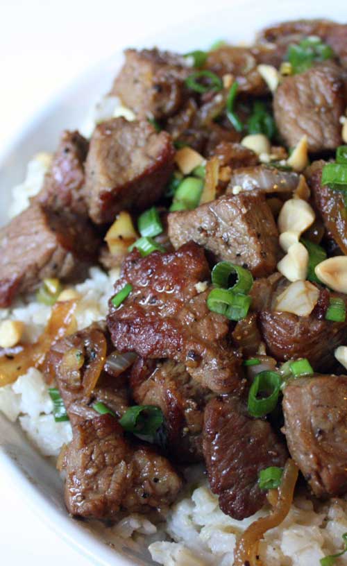 Asian-Style Garlic Beef served over white rice, on a white plate. The meat is garnished with scallions and finely chopped peanuts.