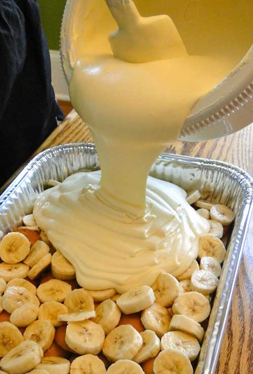 Banana pudding being poured over banana slices in a foil pan.