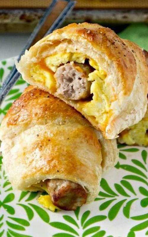 Two Sausage, Egg and Cheese Breakfast Roll-Ups on a white and green floral plate. One of the roll-ups is split open to show the inside.