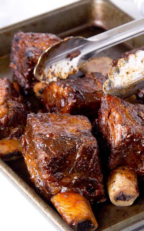 Five short ribs in a metal pan. A pair of silver tongs is preparing to pick u pone of the ribs.